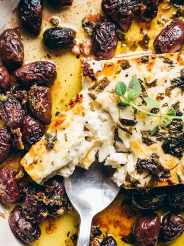 A baked feta cheese block topped with herbs, surrounded by roasted olives on a plate. A spoon is partially scooping the feta.