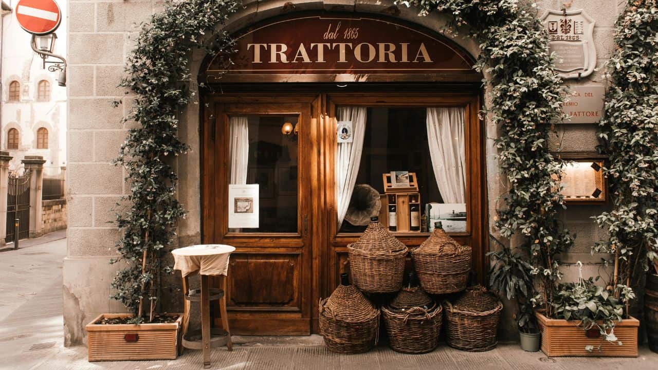 A rustic trattoria entrance with wooden door, ivy-covered facade, and wicker baskets placed outside. A small table with a white cloth is nearby.