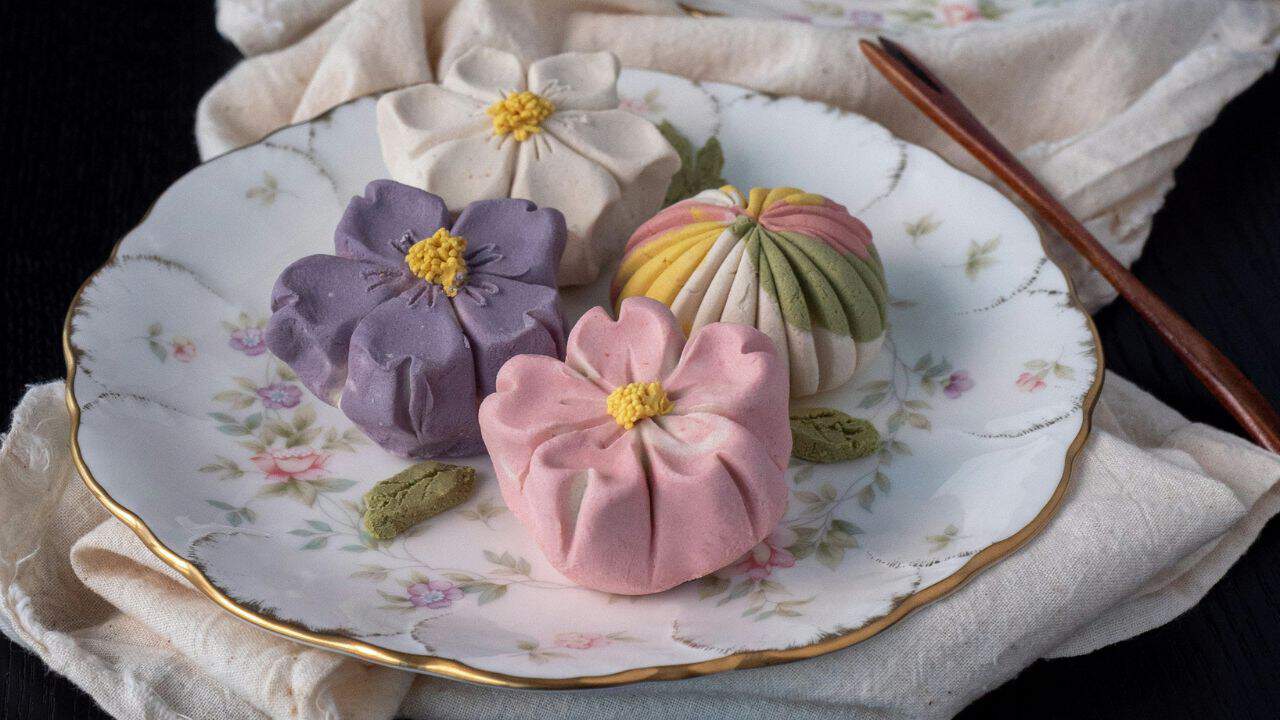 A decorative plate holds four colorful, flower-shaped confections with folded petal designs. A wooden utensil lies beside the plate on a fabric surface.