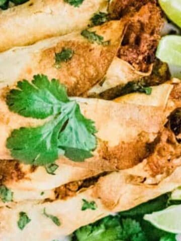 A plate of crispy rolled tacos filled with meat, garnished with fresh cilantro, accompanied by lime wedges on the side.