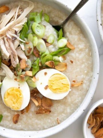 Bowl of porridge with shredded chicken, sliced boiled egg, lime wedges, green onions, and crispy garlic.