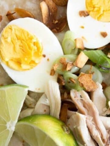 A bowl of congee topped with sliced boiled egg, shredded chicken, sliced green onions, garlic chips, and lime wedges.