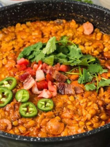 A black pot filled with lentil stew, topped with sliced jalapeños, diced tomatoes, bacon, and cilantro.