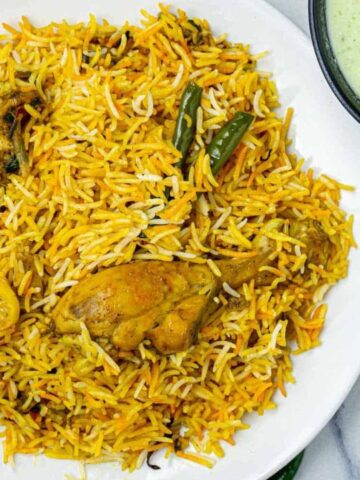 A plate of yellow chicken biryani garnished with green chilies and star anise, served alongside white yogurt sauce with cilantro. Two garlic cloves and a cloth are nearby.