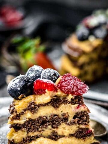 A slice of chocolate layer cake with creamy frosting, topped with blueberries and raspberries, is placed on a plate, with the rest of the cake in the background.