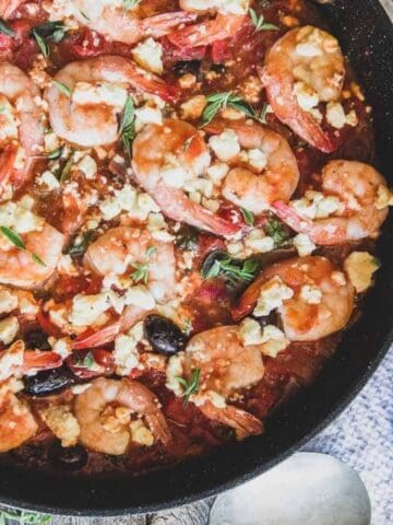 Skillet with cooked shrimp, tomatoes, olives, and crumbled cheese. A slice of rustic bread and a bag labeled "Shrimp Extra Large" are nearby.