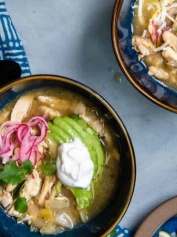 Two bowls of stew garnished with sliced avocado, pink onions, cilantro, and sour cream. Blue-patterned napkin and black utensils on the side. Slice of avocado on a small plate nearby.