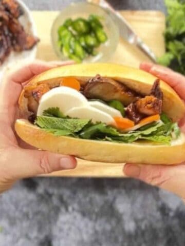 A person holds a sandwich filled with sliced meat, vegetables, and greens. A cutting board with ingredients is in the background.