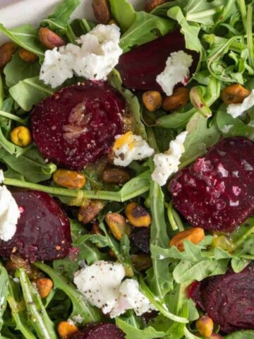 Salad with arugula, roasted beets, pistachios, and goat cheese crumbles on a white platter.