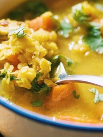 A bowl of yellow lentil soup with vegetables and cilantro, with a spoon resting in the bowl.