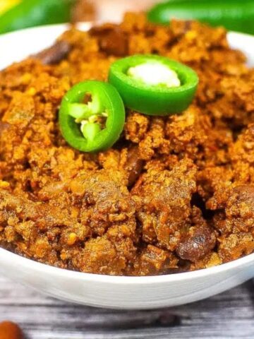 A bowl of chili con carne topped with sliced green jalapeños, with a spoon and blue-striped cloth nearby.