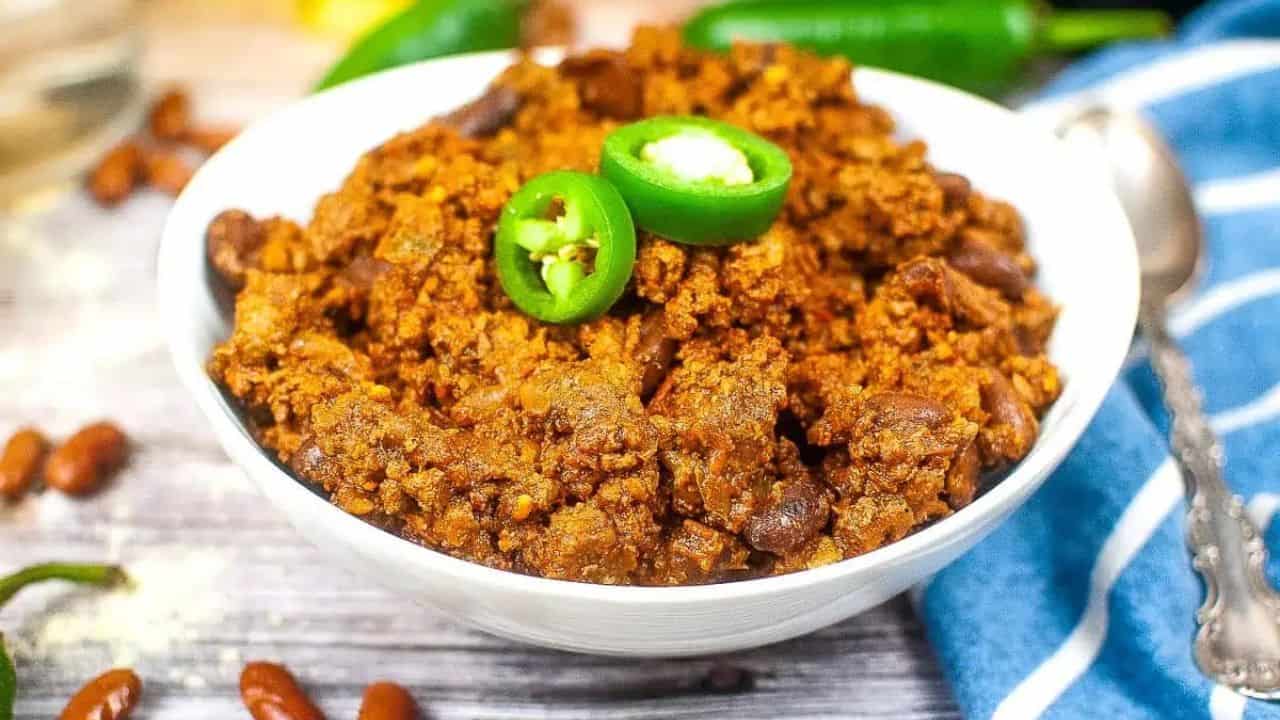 A bowl of chili con carne topped with sliced green jalapeños, with a spoon and blue-striped cloth nearby.