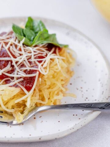 A plate of spaghetti squash topped with tomato sauce, grated cheese, and basil, with a fork beside it.
