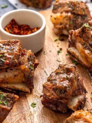 Grilled marinated beef ribs garnished with herbs on a wooden board, served with a small bowl of red pepper flakes.