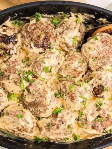 A black dish filled with meatballs covered in creamy sauce, garnished with chopped parsley, and a wooden spoon resting on the side.