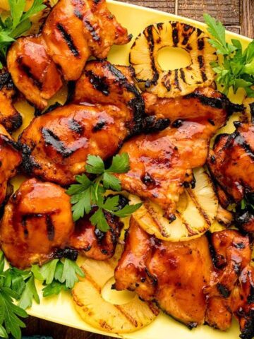 Grilled chicken with barbecue glaze and pineapple slices, garnished with parsley, served on a yellow platter.
