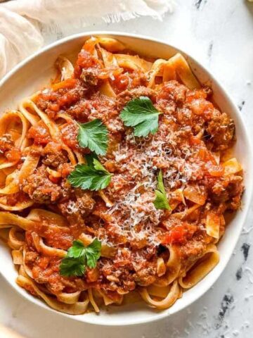 A plate of pasta topped with meat sauce and garnished with parsley and grated cheese, accompanied by a piece of crusty bread on a marble surface.