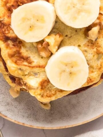 A plate of banana pancakes topped with banana slices and walnuts. An egg and cutlery are nearby on a white tabletop.