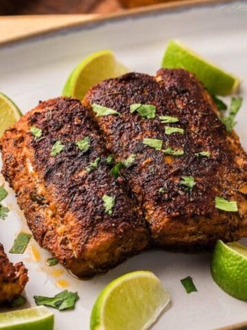 Seasoned blackened fish fillets garnished with chopped herbs and surrounded by lime wedges on a white rectangular plate.