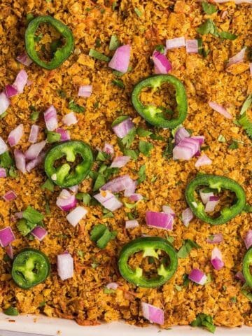A casserole dish with a golden-brown topping, garnished with sliced jalapeños, chopped red onions, and green herbs.