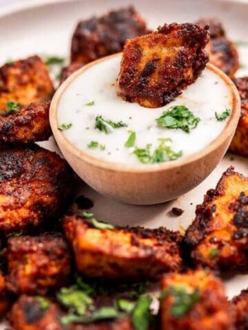 Spicy grilled chicken bites garnished with cilantro, served around a bowl of creamy white sauce on a white plate.
