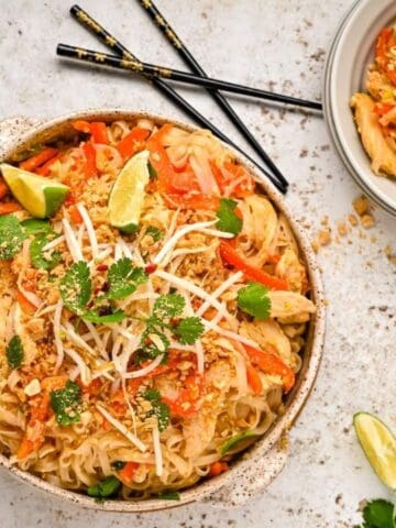 Two bowls of Pad Thai garnished with lime wedges, cilantro, and bean sprouts. Chopsticks and herbs are scattered around on a light surface.