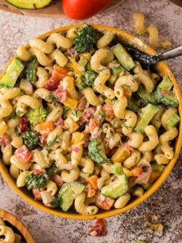 A large bowl of pasta salad with broccoli, avocado, tomatoes, and herbs, surrounded by smaller bowls of similar salad on a textured surface. A yellow napkin is nearby.
