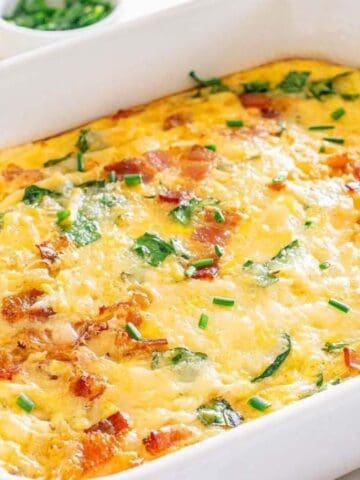 Baked casserole in a white dish, featuring golden cheese, green herbs, and visible bacon pieces. White plates and ingredients are nearby.