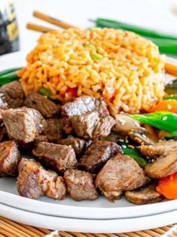A plate with beef chunks, fried rice, and stir-fried vegetables, placed on a bamboo mat. Soy sauce bottle and green onions in the background.