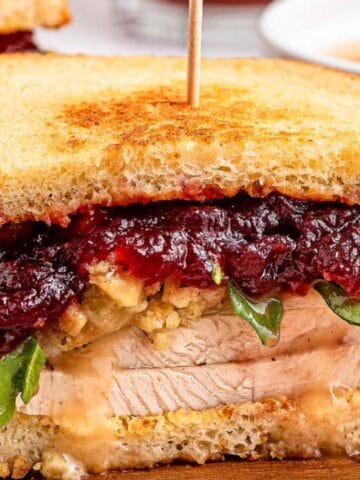 Turkey sandwich with stuffing, arugula, cheese, cranberry sauce on toasted bread, and a wooden pick. Served with chips and a small bowl of gravy in the background.