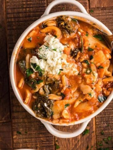 A bowl of tomato-based pasta stew with greens, topped with ricotta and herbs, on a wooden table. A spoon is placed inside the bowl.