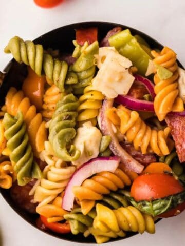 A bowl of colorful rotini pasta salad with tomatoes, cheese, red onion, and peppers on a white surface.