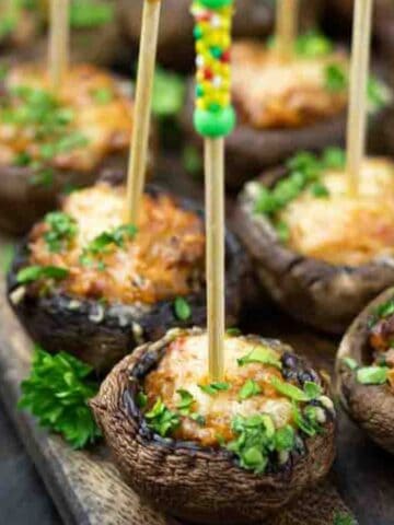 Stuffed mushrooms topped with cheese and herbs, each held by a skewer with colorful beads, arranged on a wooden tray.