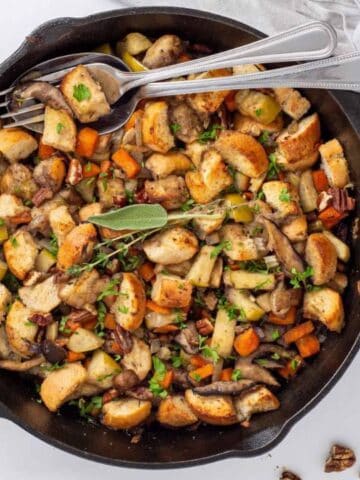 A skillet filled with a stuffing mixture containing bread cubes, mushrooms, carrots, and herbs. Several pieces of cutlery rest on a napkin beside the skillet.