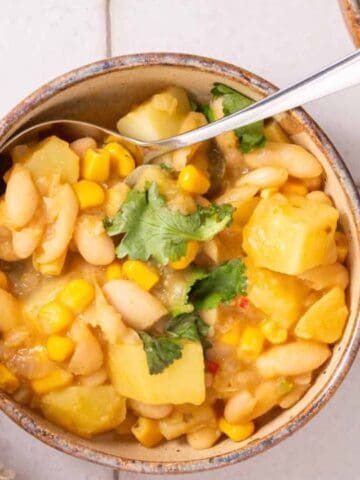 A bowl of white bean and corn soup with potatoes and cilantro garnish, placed on a tiled surface with a spoon in the bowl.