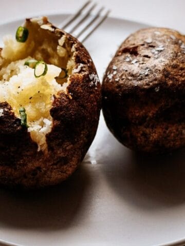 Two baked potatoes on a white plate; one is split open with butter and green onions, accompanied by a fork in the background.