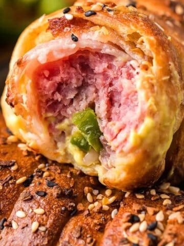 Close-up of a sliced savory pastry roll filled with ham, cheese, and green peppers, topped with sesame seeds, on a black plate.