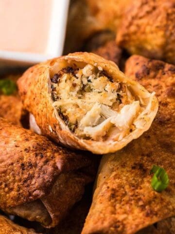Close-up of crispy egg rolls filled with chicken and cheese, garnished with chopped green onions. A dipping sauce is in a white bowl in the background.