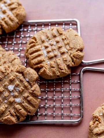 Peanut butter cookies with fork marks and sea salt on a cooling rack, with a spoonful of peanut butter in the background.