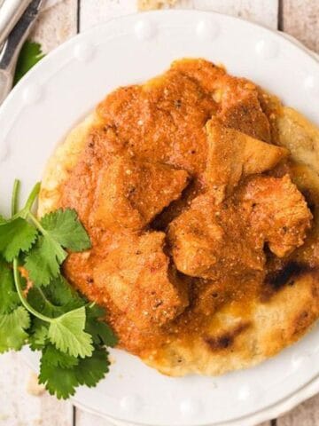 Plate of chicken curry on naan bread, garnished with cilantro. Fork and knife beside the plate on a wooden table.