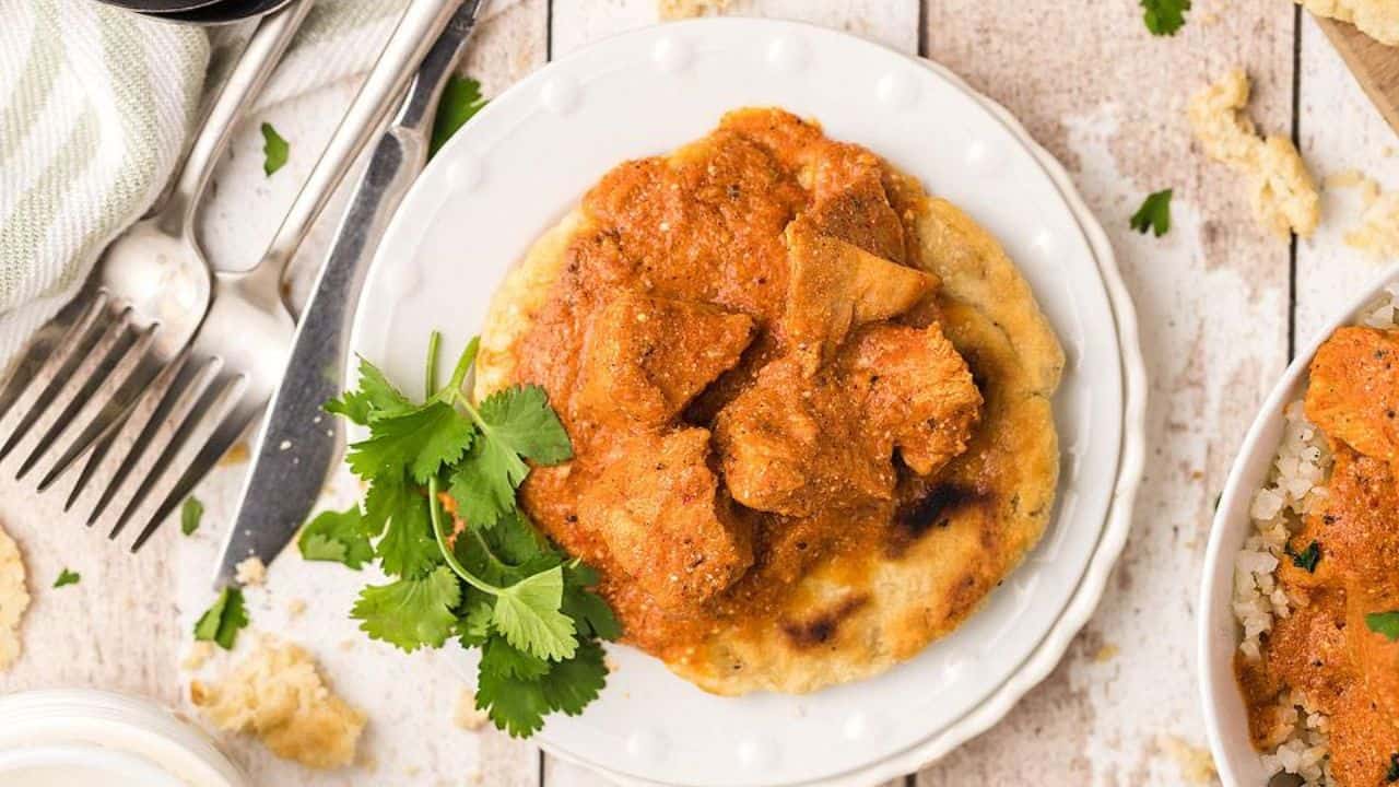 Plate of chicken curry on naan bread, garnished with cilantro. Fork and knife beside the plate on a wooden table.