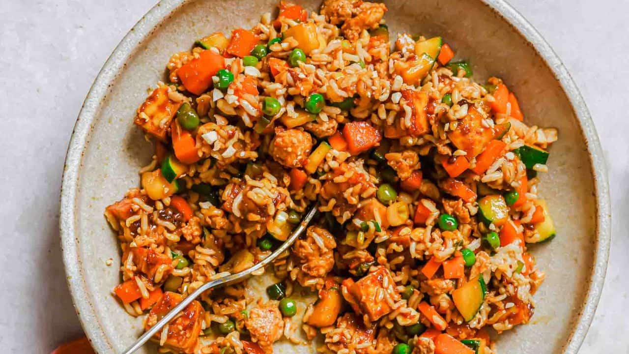 A bowl of fried rice with vegetables and pieces of meat, featuring carrots, peas, and zucchini, with a spoon.
