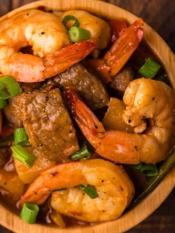 A bowl of stew with shrimp, beef, pineapple chunks, and chopped green onions, placed on a wooden surface.