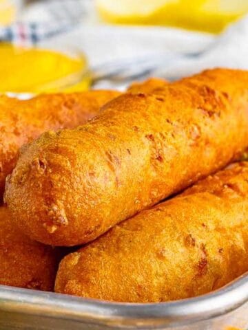 Three corn dogs stacked in a metal tray, with a side of leafy greens nearby.
