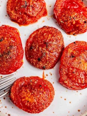 A white plate with six sliced and seasoned roasted tomatoes, a fork placed nearby, on a wooden surface with a striped cloth.
