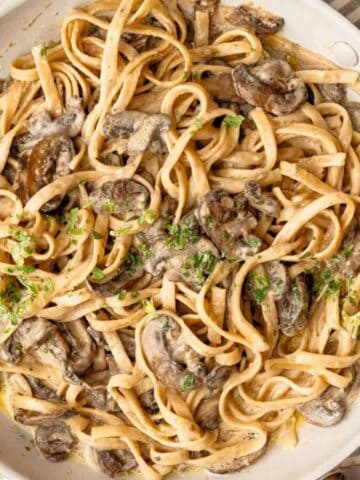 A pan of fettuccine pasta with mushrooms and cream sauce, garnished with parsley, placed on a striped cloth.