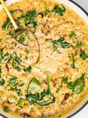 A pot of creamy pasta with spinach and sun-dried tomatoes on a marble surface, accompanied by a cheese grater, parsley, and salt and pepper shakers.