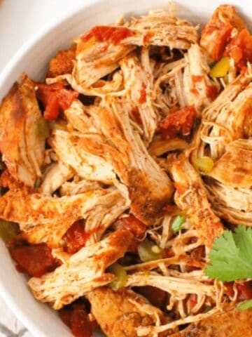 A bowl of shredded chicken mixed with tomatoes and spices, garnished with fresh cilantro. Sliced lime and chopped red onion are on a wooden board nearby.