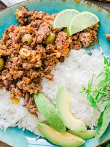 A colorful plate of rice topped with seasoned ground meat, green olives, avocado slices, lime wedges, and fresh herbs on a wooden table.