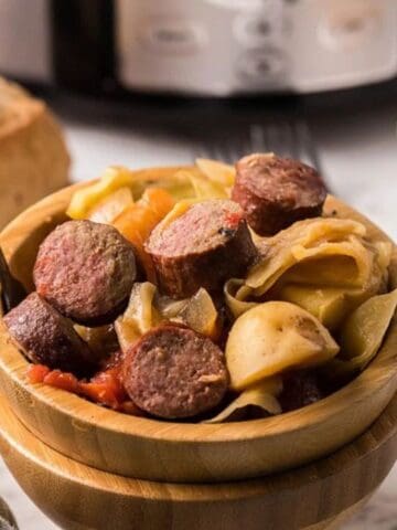 A bowl of sliced sausage with vegetables and sliced potatoes. Rolls are in the background, and a fork is placed beside the bowl.